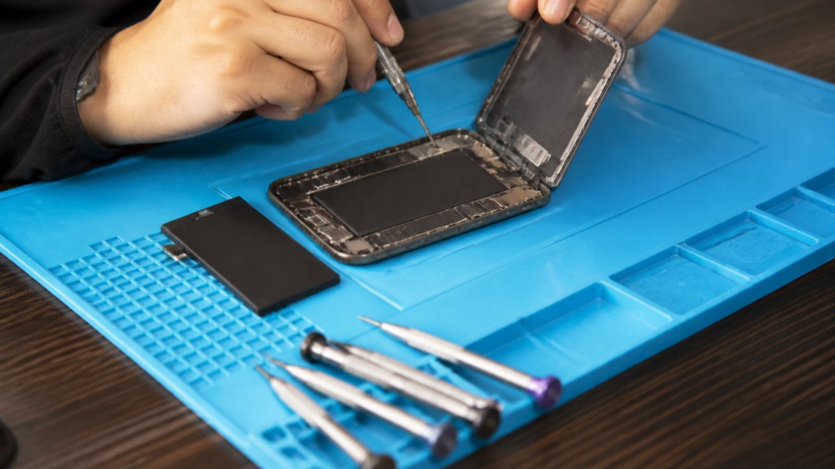 A repair technician at a clean workbench using precision tools to open an iPhone, with a replacement battery and screwdrivers laid out nearby